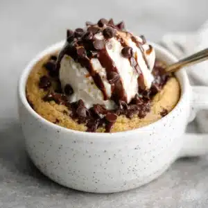 A warm high protein mug cake in a white mug, topped with vanilla ice cream, melted chocolate drizzle, and mini chocolate chips