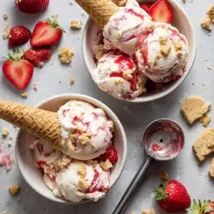 No-churn strawberry shortcake ice cream with fresh strawberries and shortcake chunks in a bowl, styled with cone and toppings for National Ice Cream Day