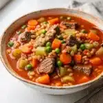 Hearty beef and barley soup with carrots and peas in a rustic ceramic bowl.